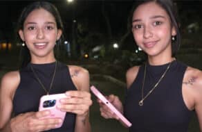 Camila Cooper en la calle de noche con top negro y collar sonriendo a cámara mientras sostiene un teléfono rosa